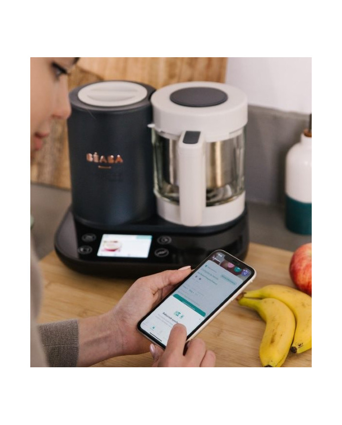 Mama naudoja Béaba ir Me programėlę telefone šalia Babycook Smart prietaiso virtuvėje