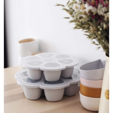 Dvi sustatytos BEABA Multiportions XL silikoninės formos ant medinio stalo šalia dubenėlių ir gėlių puokštės